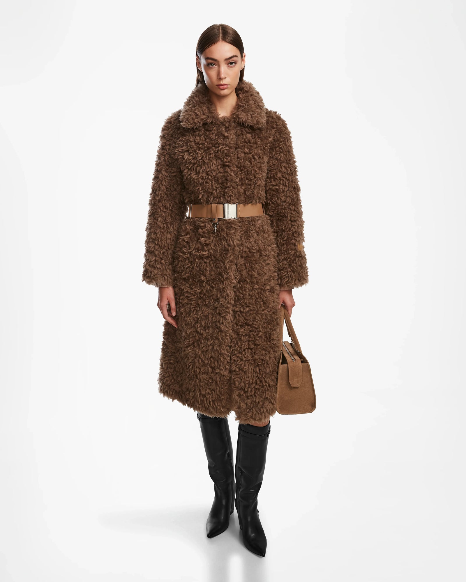 A woman faces forward against a white background wearing the Rudsak KAMILAH - 8124827 Camel faux fur trench with a vegan leather belt, black knee-high boots, and holding a matching brown handbag.