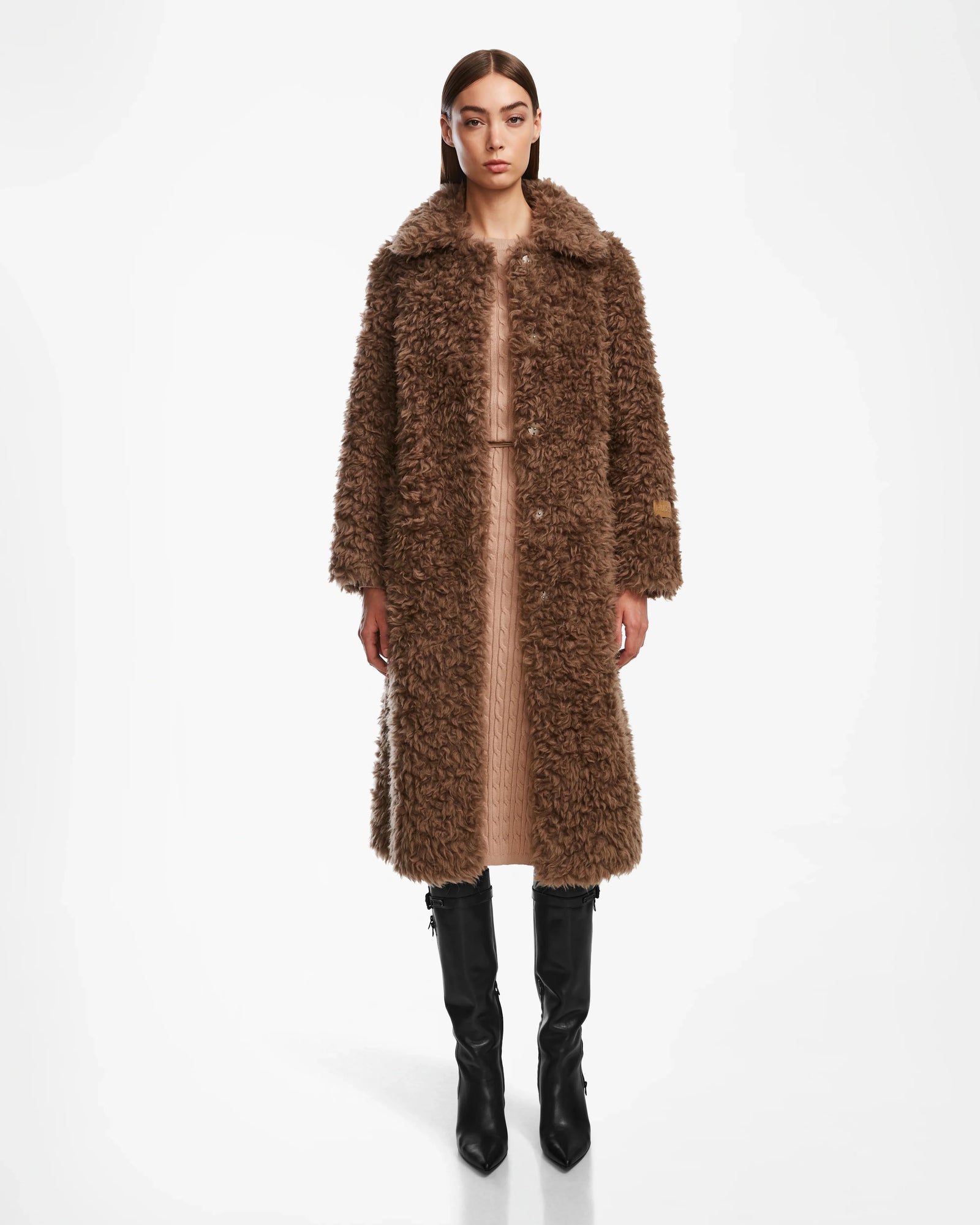 A woman stands against a white background wearing the Rudsak KAMILAH - 8124827 Camel faux fur trench coat over a beige dress and knee-high black boots, looking straight ahead with her arms relaxed by her sides.