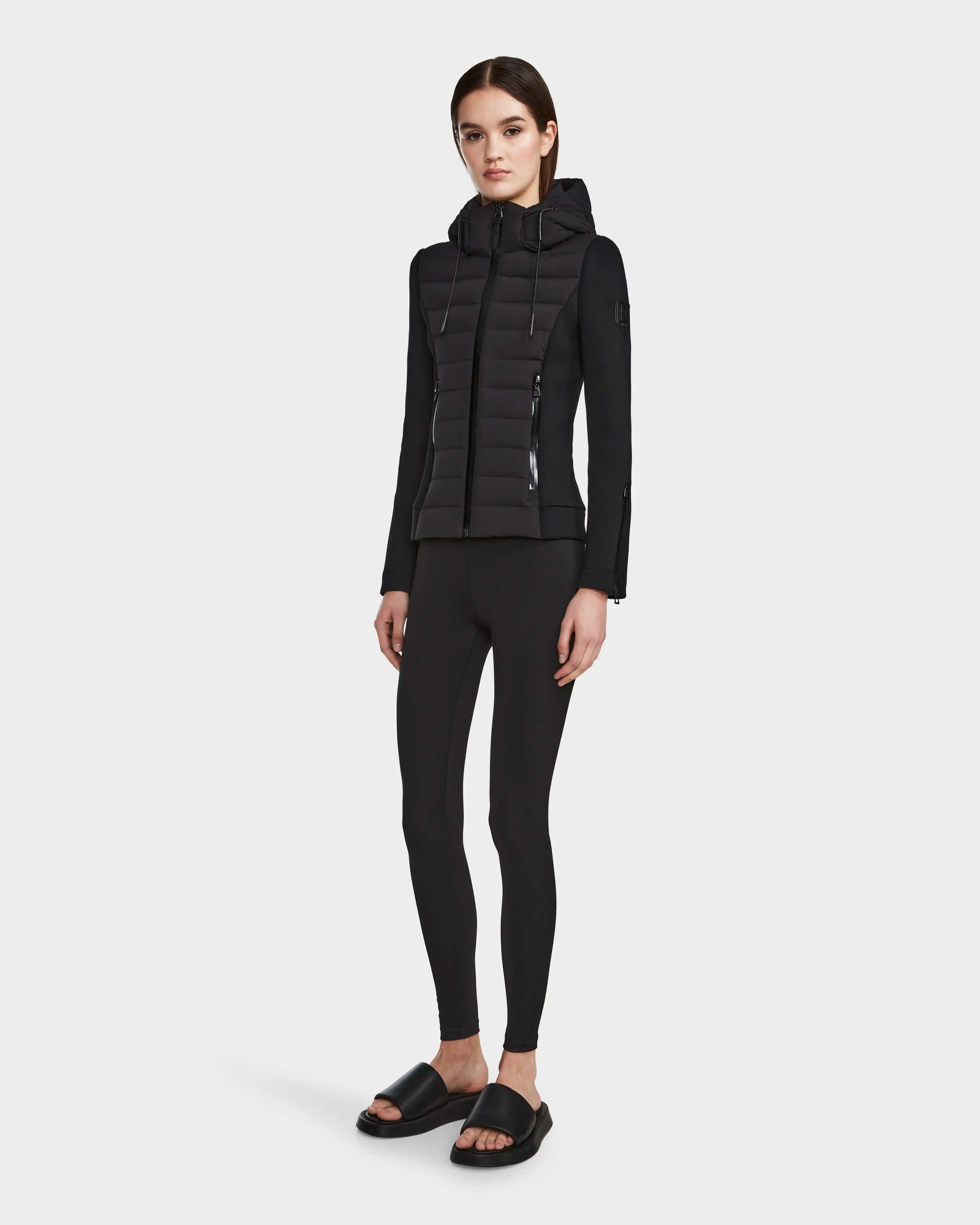 A woman with straight brown hair and a neutral expression stands facing forward, wearing the RUDSAK MABS - 8122553 BLACK lightweight activewear jacket, black leggings, and black slide sandals against a plain white background.