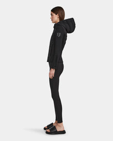 A woman stands in profile against a white background, wearing the RUDSAK MABS - 8122553 BLACK women’s puffer jacket with RDS-certified down, paired with black leggings and black slide sandals.