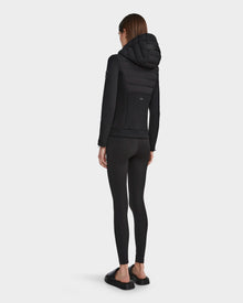 A woman faces away, wearing the RUDSAK MABS - 8122553 BLACK lightweight activewear jacket with black leggings and slide sandals against a plain white background.