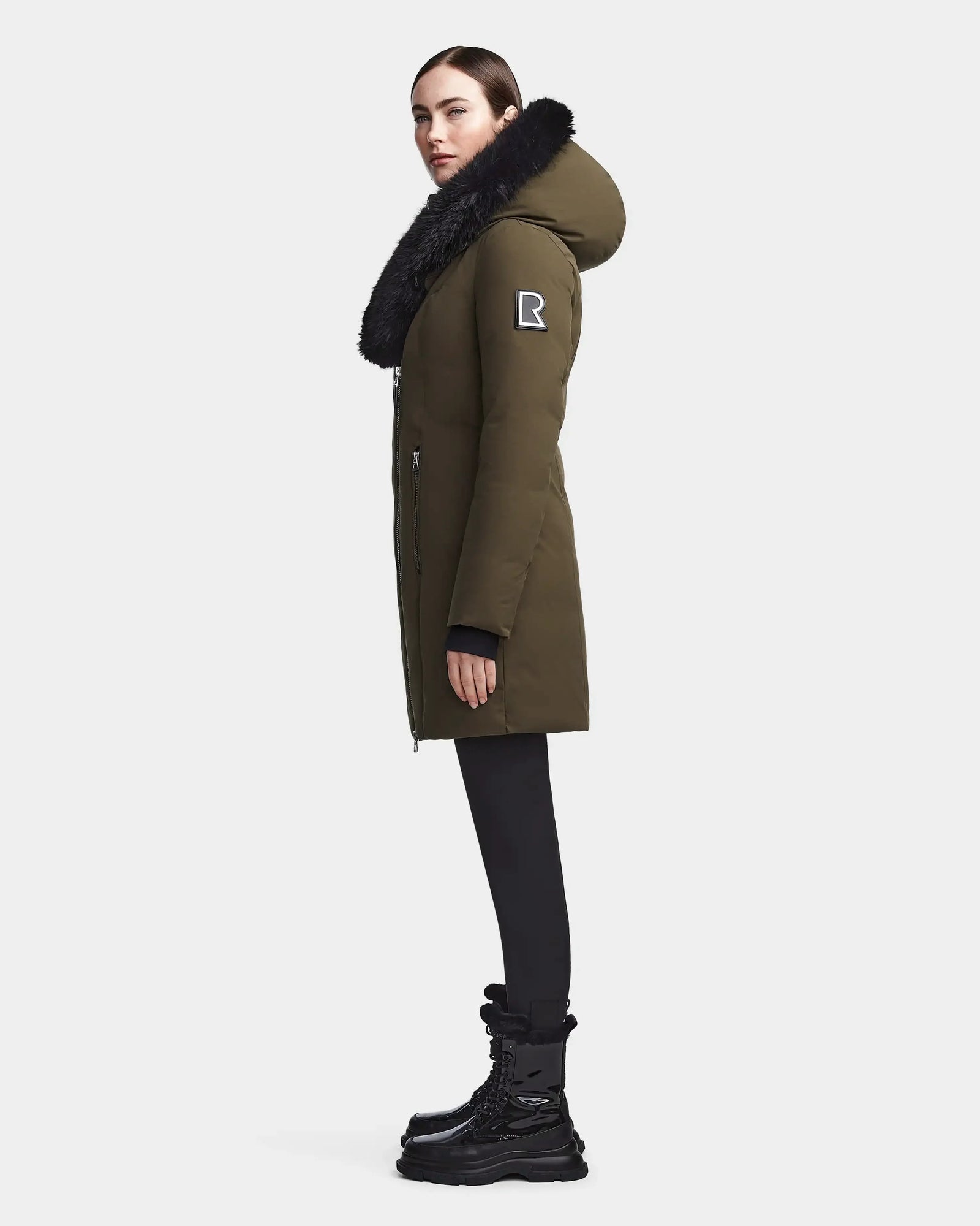 A woman stands in profile wearing a Rudsak MAIA M - 8123582 Army, an olive green knee-length women’s down parka with a large black fur-trimmed hood and R badge on the sleeve, paired with black leggings and winter boots.