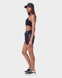 A woman stands sideways wearing the Rudsak RYLEE - 8523001 BLACK running shorts with spandex inner short, paired with a black sports bra, cap, and black-and-white sneakers against a plain white background.