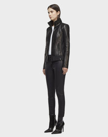 A woman with straight dark hair stands against a plain white background, wearing the Rudsak PRESLEY - CA EN 8119029 BLACK biker jacket, styled with black pants, a white top, and black heeled ankle boots.