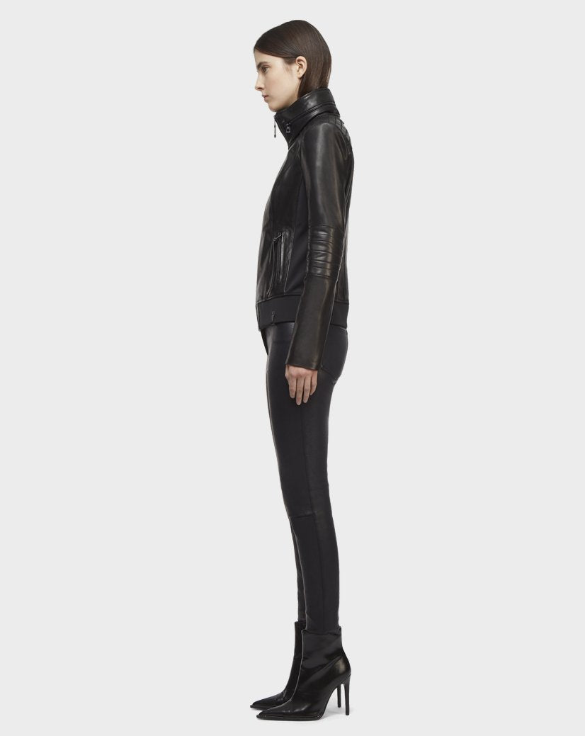 A person with straight brown hair models the Rudsak PRESLEY - CA EN 8119029 BLACK lamb leather biker jacket, black skinny pants, and black heeled boots, standing in profile facing left against a plain white background.