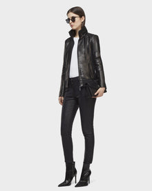 A woman in Rudsak’s PRESLEY - CA EN 8119029 BLACK lamb leather biker jacket, sunglasses, white top, black leather pants, and heeled boots holds a black clutch against a plain white background.