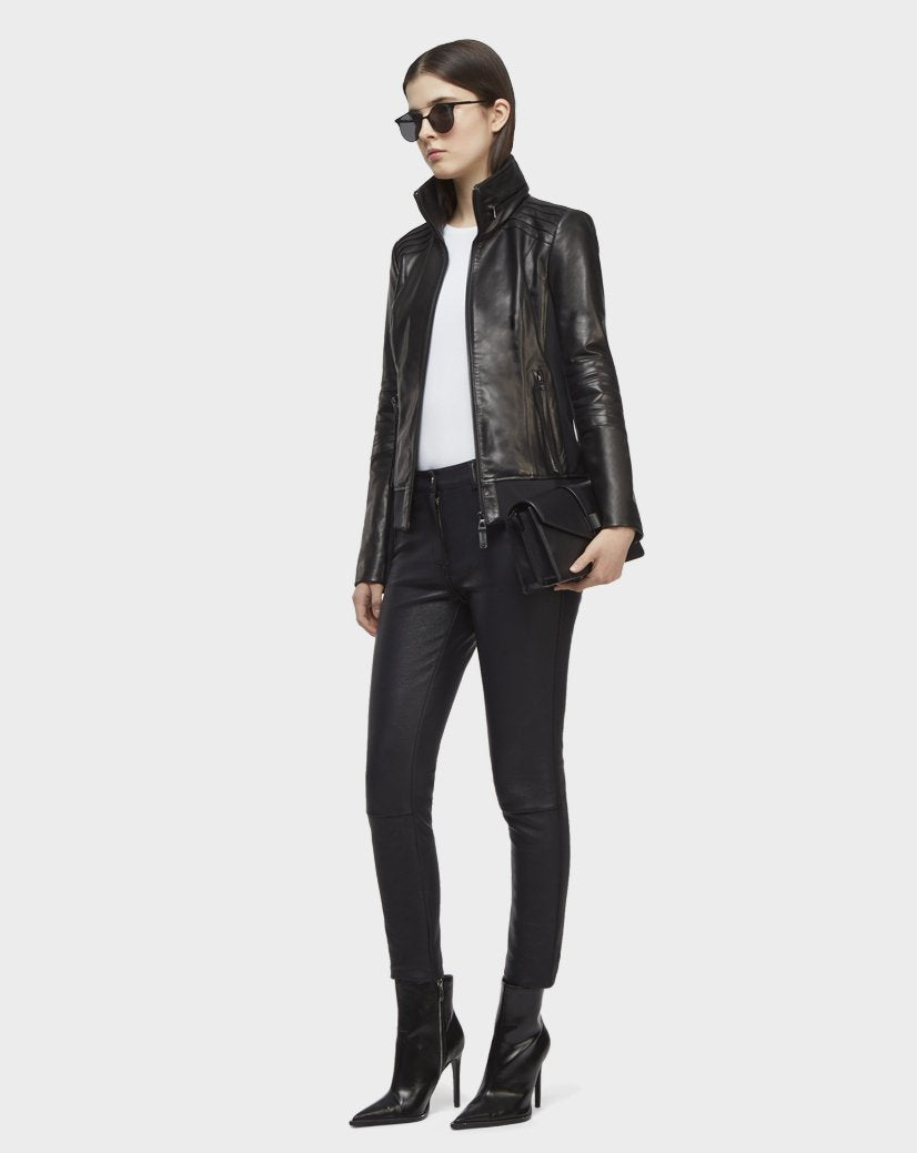 A woman in Rudsak’s PRESLEY - CA EN 8119029 BLACK lamb leather biker jacket, sunglasses, white top, black leather pants, and heeled boots holds a black clutch against a plain white background.