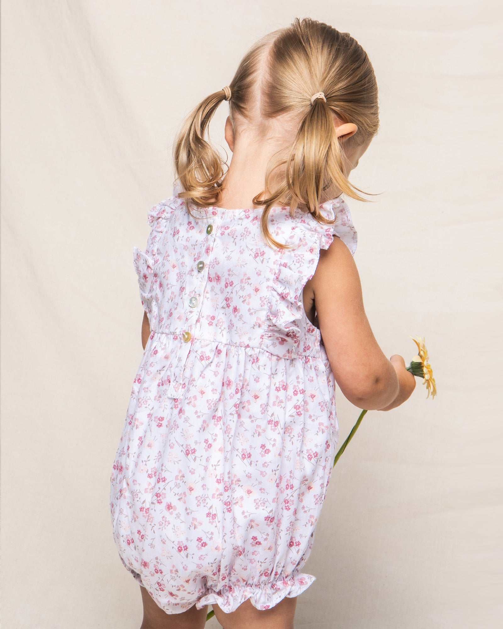 A young girl with pigtails, dressed in Petite Plumes Babys Twill Ruffled Romper in Dorset Floral, is turned away holding a sunflower against a plain light-colored background.