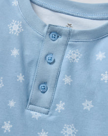 The Kid's Pima Snug Fit Pajama Set in Periwinkle Wonderland features light blue, long-sleeve pajamas with white snowflakes, buttoned neckline, matching elastic waist pants, and cuffed ankles. Shown laid flat on a white background.