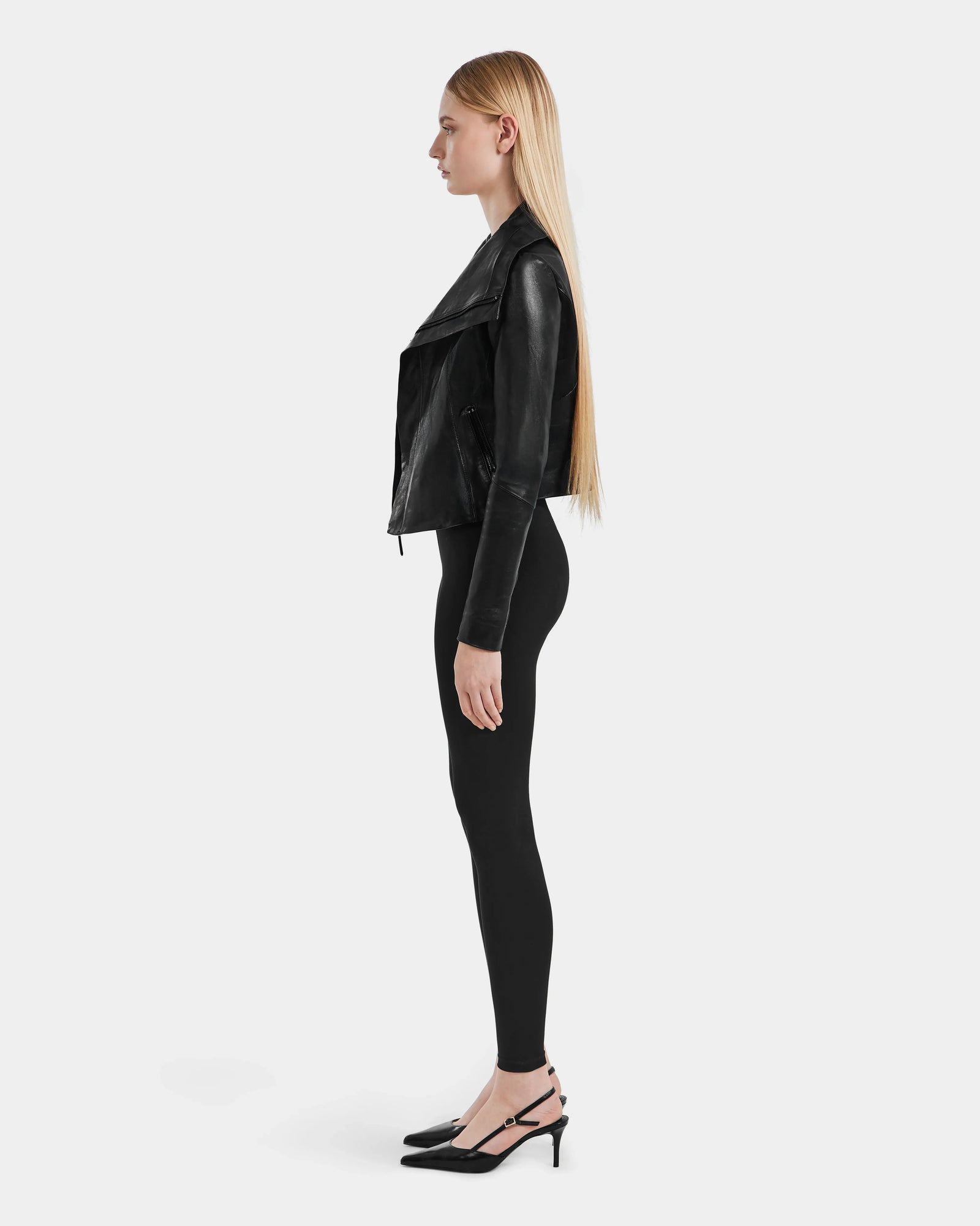 A woman with long straight blonde hair stands in profile against a plain background, wearing the Rudsak AVRIL - 8124561 Black jacket, black leggings, and black heeled shoes.