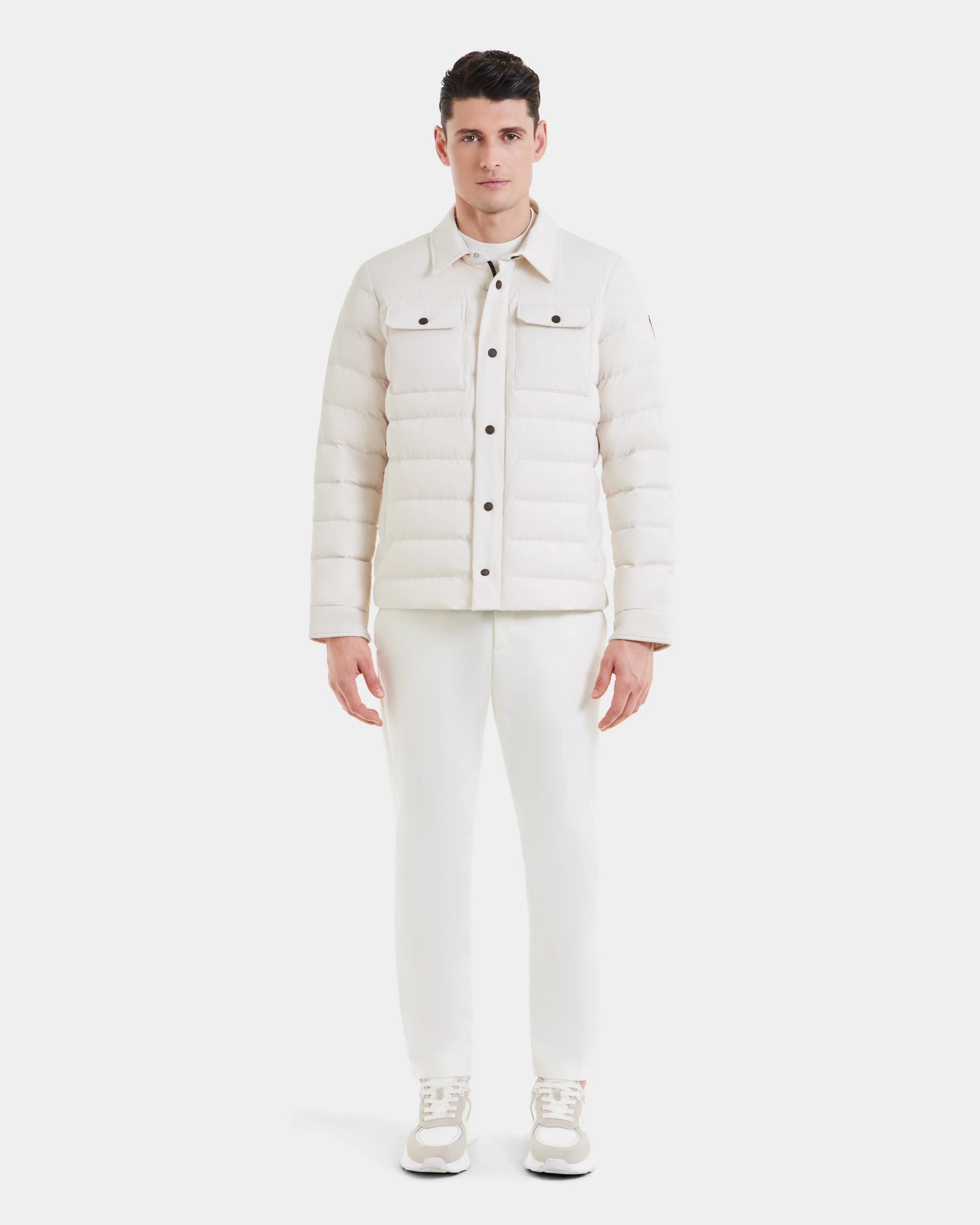 A man faces forward against a plain white background, wearing the Rudsak AZIEL - M124521 Vanilla duck down jacket with matching white pants and sneakers.