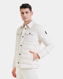 A man wears the Rudsak AZIEL - M124521 Vanilla quilted mens puffer shirt jacket with black buttons and flap pockets, standing with one hand in his pocket against a plain white background.