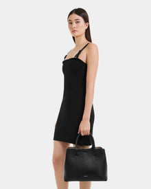 A woman in a sleeveless black dress holds the Rudsak CATIRA S - 8325006 Black leather tote by her side. She has long, straight dark hair and stands before a plain white background.
