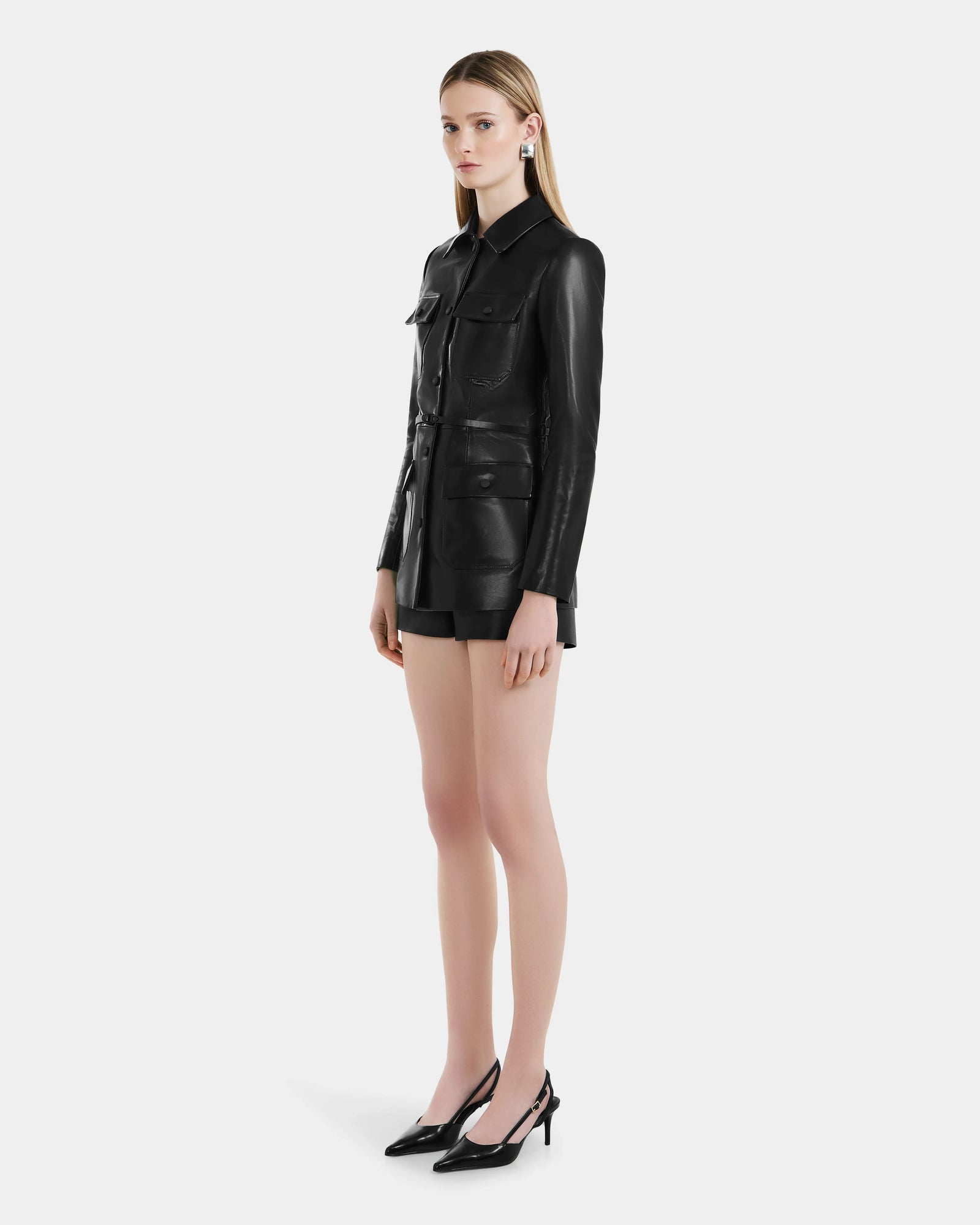 A woman with long blonde hair and a neutral expression wears the Rudsak DANICA - 8125025 Black womens leather jacket, styled with front pockets, black shorts, and black high heels against a plain white background.