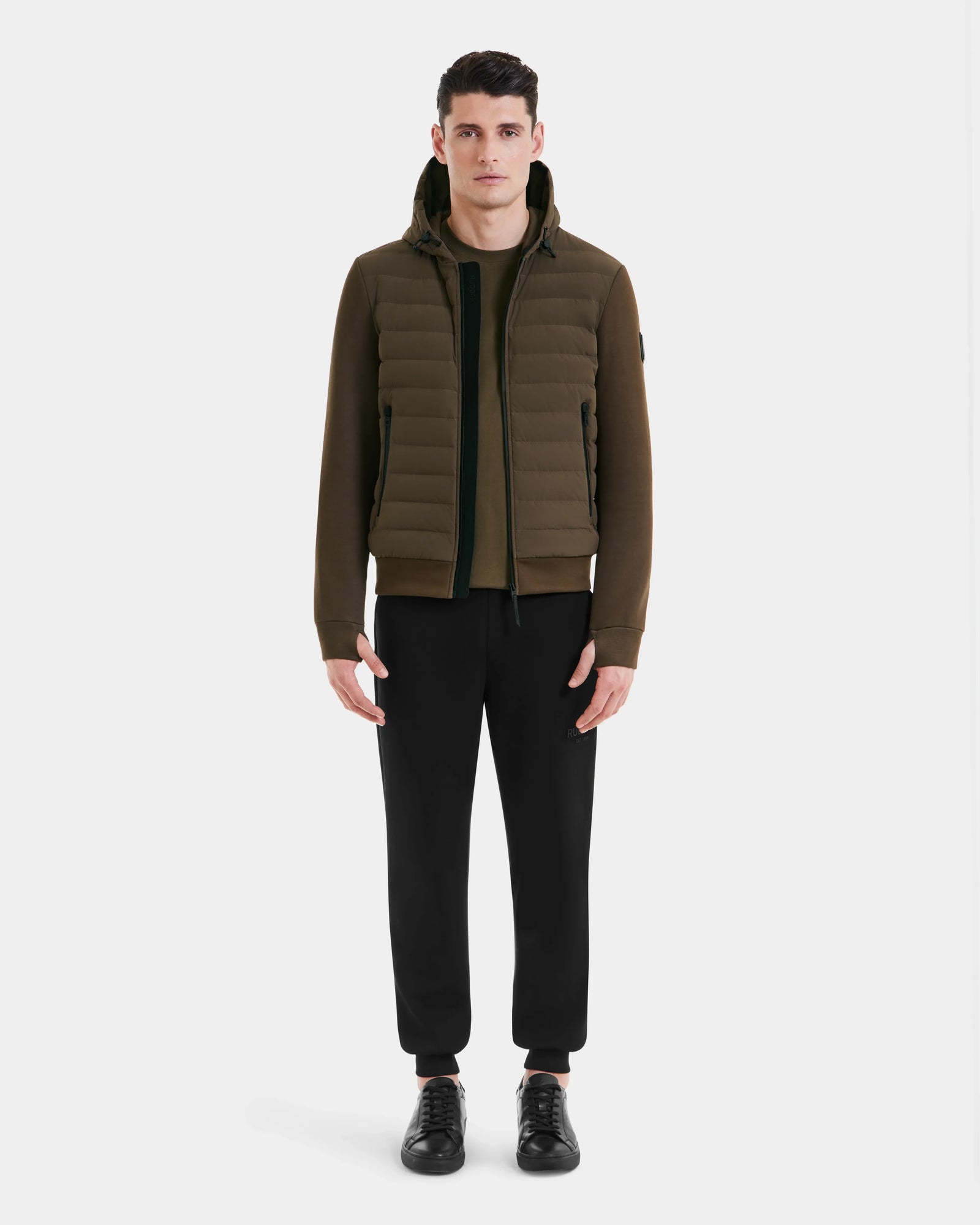 A man faces forward against a white background, wearing the Rudsak DEE MAT - 6124028 Army lightweight hybrid down jacket over a zip-up top, with black jogger pants and black sneakers.