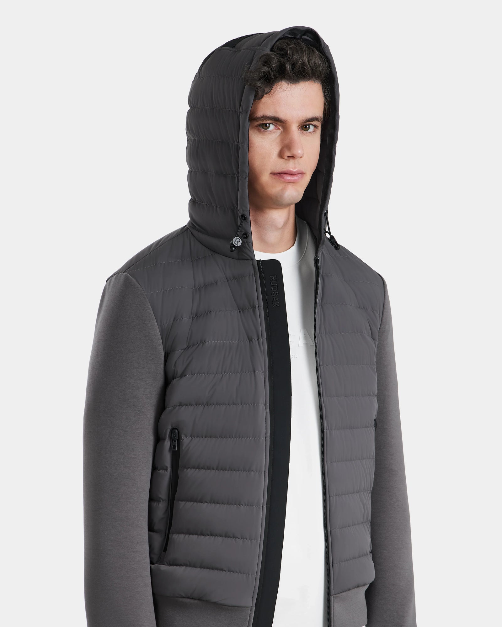 A young man with short curly hair wears the Rudsak DEE MAT - 6124028 Grey lightweight down hooded puffer over a white shirt. He looks straight ahead, hood up, zipper closed, hands in side pocket. Background is plain white.