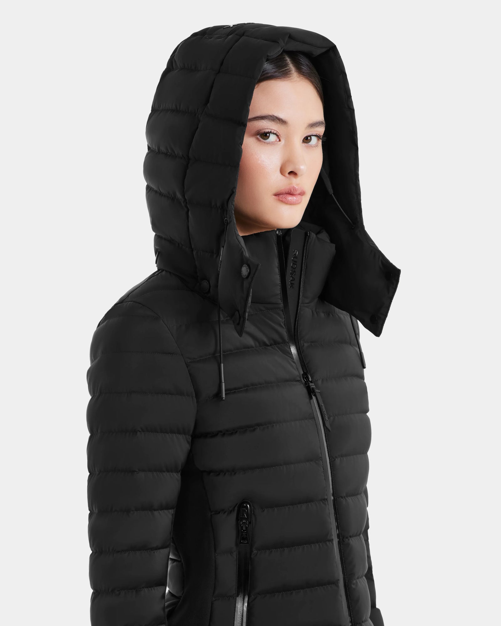 A woman stands against a plain light gray background wearing the Rudsak MARYA M - L123559 Black quilted women’s down puffer jacket with a hood, front zipper, and side pockets.