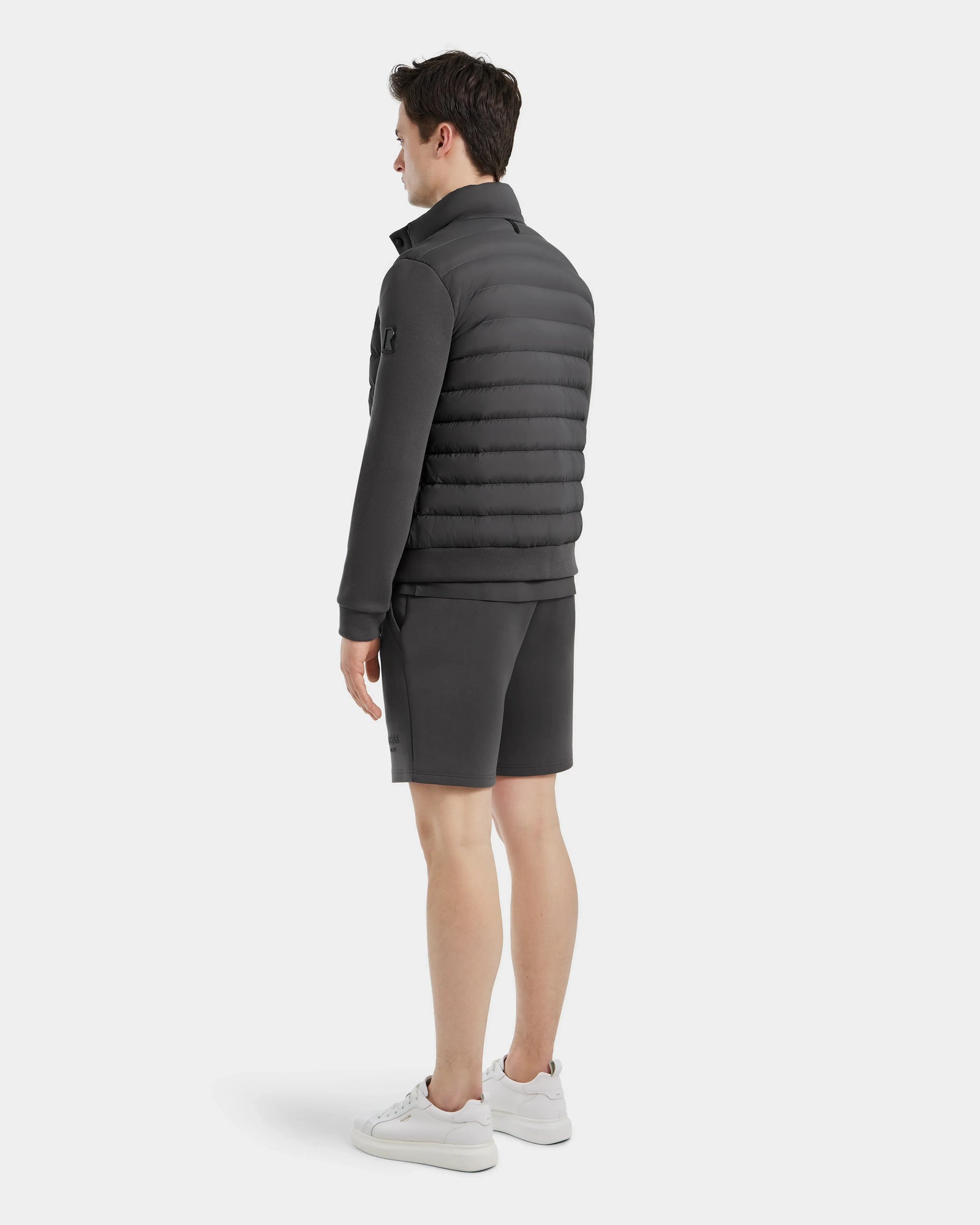 A man stands facing away against a plain light background, wearing Rudsaks RAGAB - 6124577 Grey mens hybrid jacket with lightweight down insulation, matching shorts, and white sneakers.