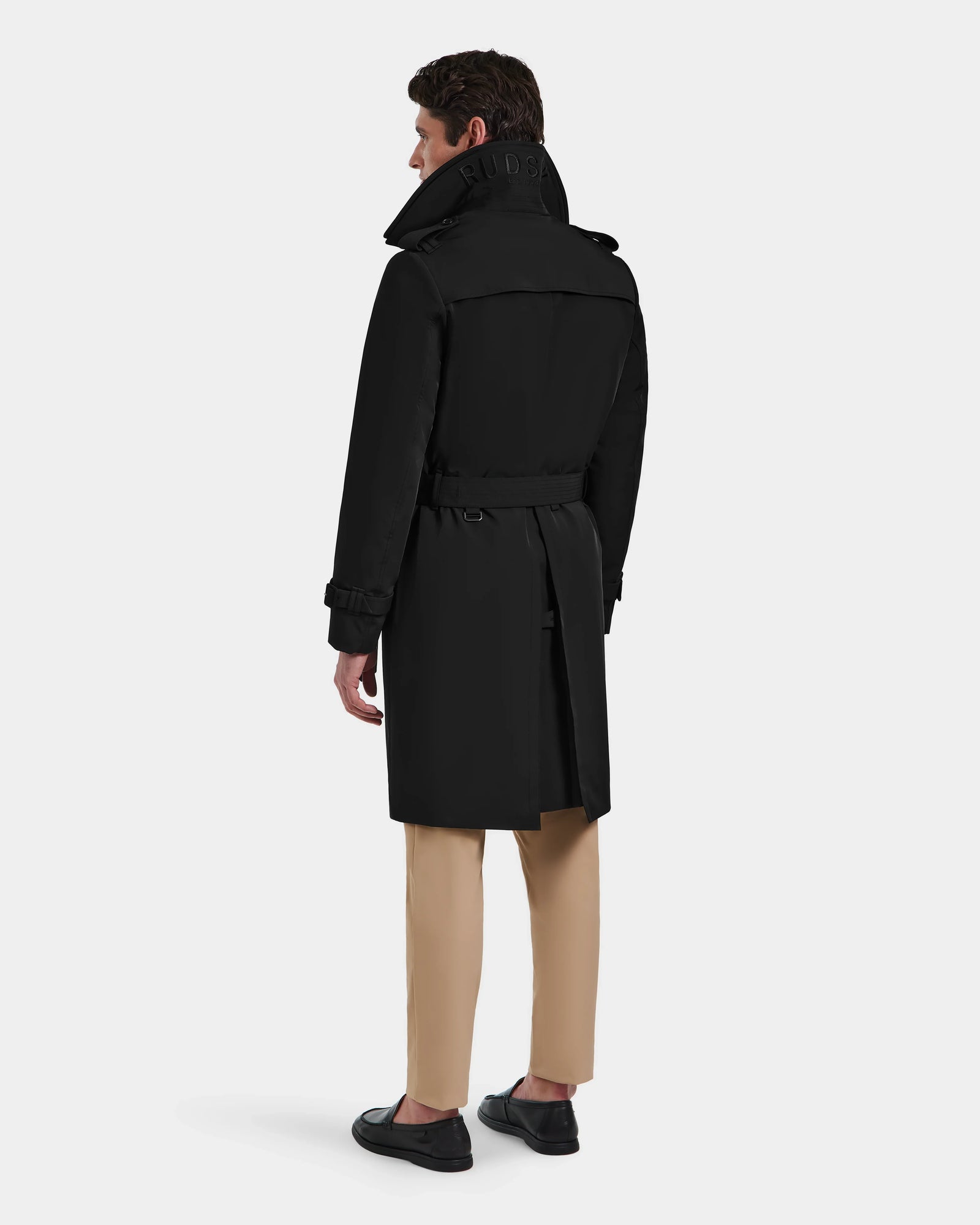 A man stands facing away, wearing the Rudsak TIMOTHY - 6124589 Black knee-length trench coat, beige pants, and black shoes against a plain light background.