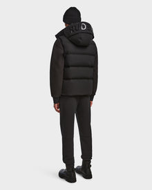 A person in the Rudsak REN M - 9123512 Black M weather-resistant puffer jacket, black beanie, pants, and shoes stands facing away from the camera against a plain light background.