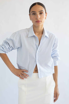 Woman wearing a light blue striped shirt and white pants against a plain background