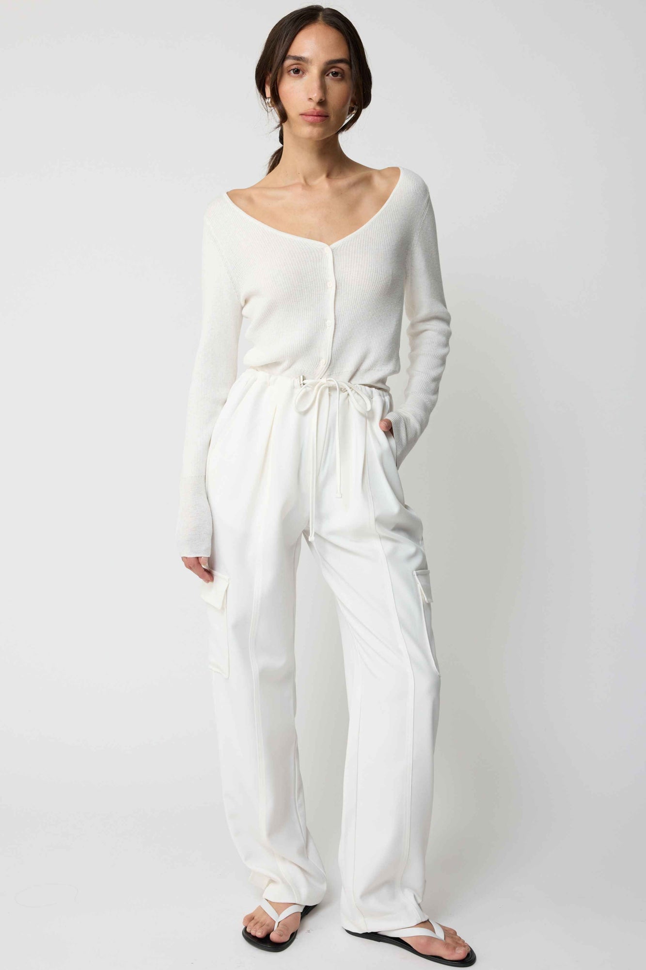 Woman wearing a white cardigan and white cargo pants on a plain background