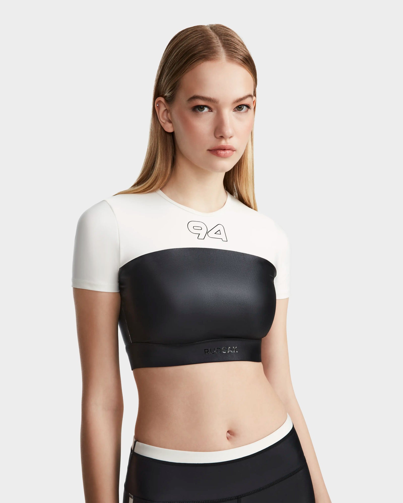 A young woman with straight, light brown hair models the Rudsak LILIANE - 8524018 Black fitted short sleeve crop top, featuring a white upper with “4A” lettering and black lower section, against a plain light gray background.