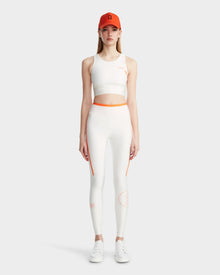 A woman faces forward wearing the Rudsak TRACY - 8524016 Eggshell quick dry sports bra and high-waisted leggings with orange accents, paired with white sneakers and an orange baseball cap. The background is light-colored and plain.