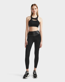 A woman faces forward wearing the RUDSAK ARYA - 8523002 BLACK sports bra with a cutout, high-waisted black leggings in MVMT+ fabric, and black sneakers, set against a plain light background.