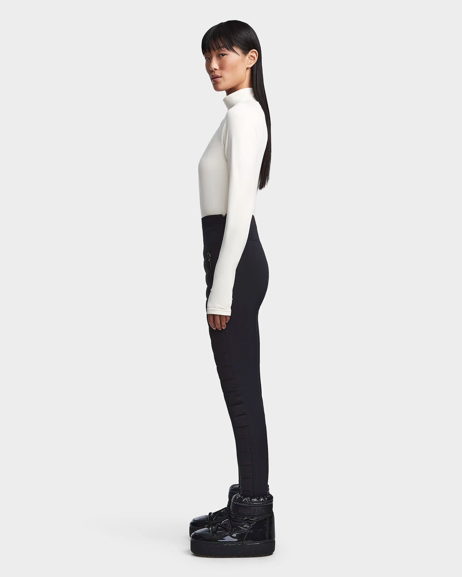 A woman in profile stands against a light gray backdrop wearing the Rudsak KILTON - 8523533 Iceland women’s fleece-lined jersey top, styled with high-waisted black pants and black platform shoes. Her long straight hair falls naturally, arms at her sides.