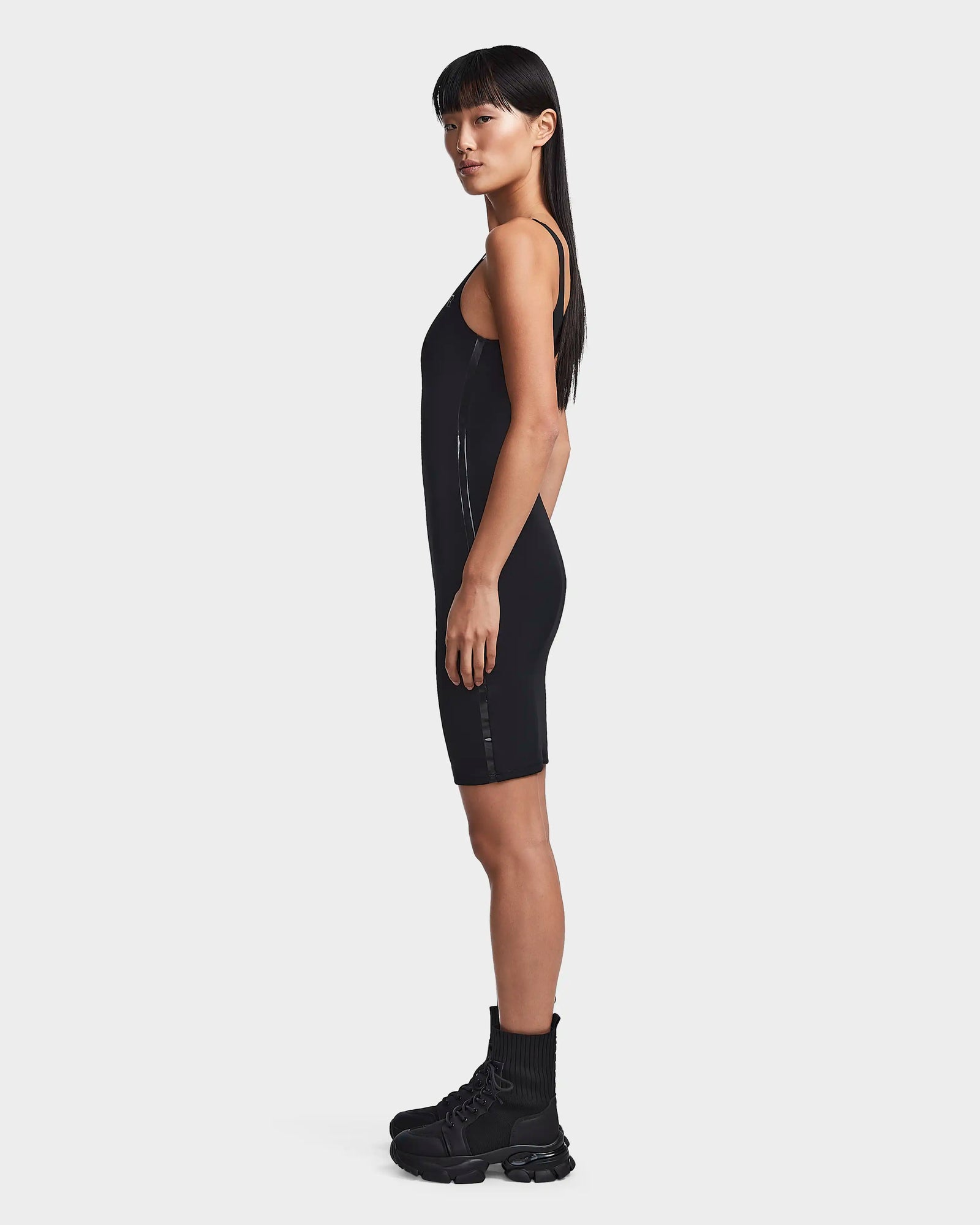 A woman in profile wears the Rudsak MILEY - 8523034 BLACK sleeveless dress with black combat boots and socks. She has long straight hair, a neutral expression, and is posed against a plain white background.
