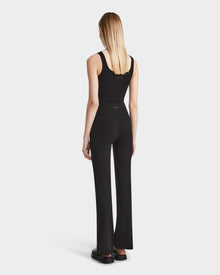 A woman with straight blonde hair stands facing away in the ADRIANA - 8524009 Black sleeveless top by Rudsak, paired with flared-leg leggings and black shoes against a plain white background.