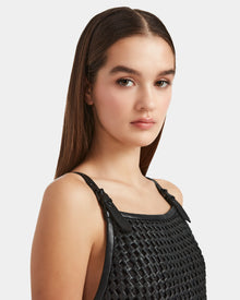 A young woman with straight brown hair models the Rudsak NATALIA - 8524045 Black dress, featuring a sleeveless, woven-texture top with buckle straps. She has a neutral expression and poses against a plain light background.