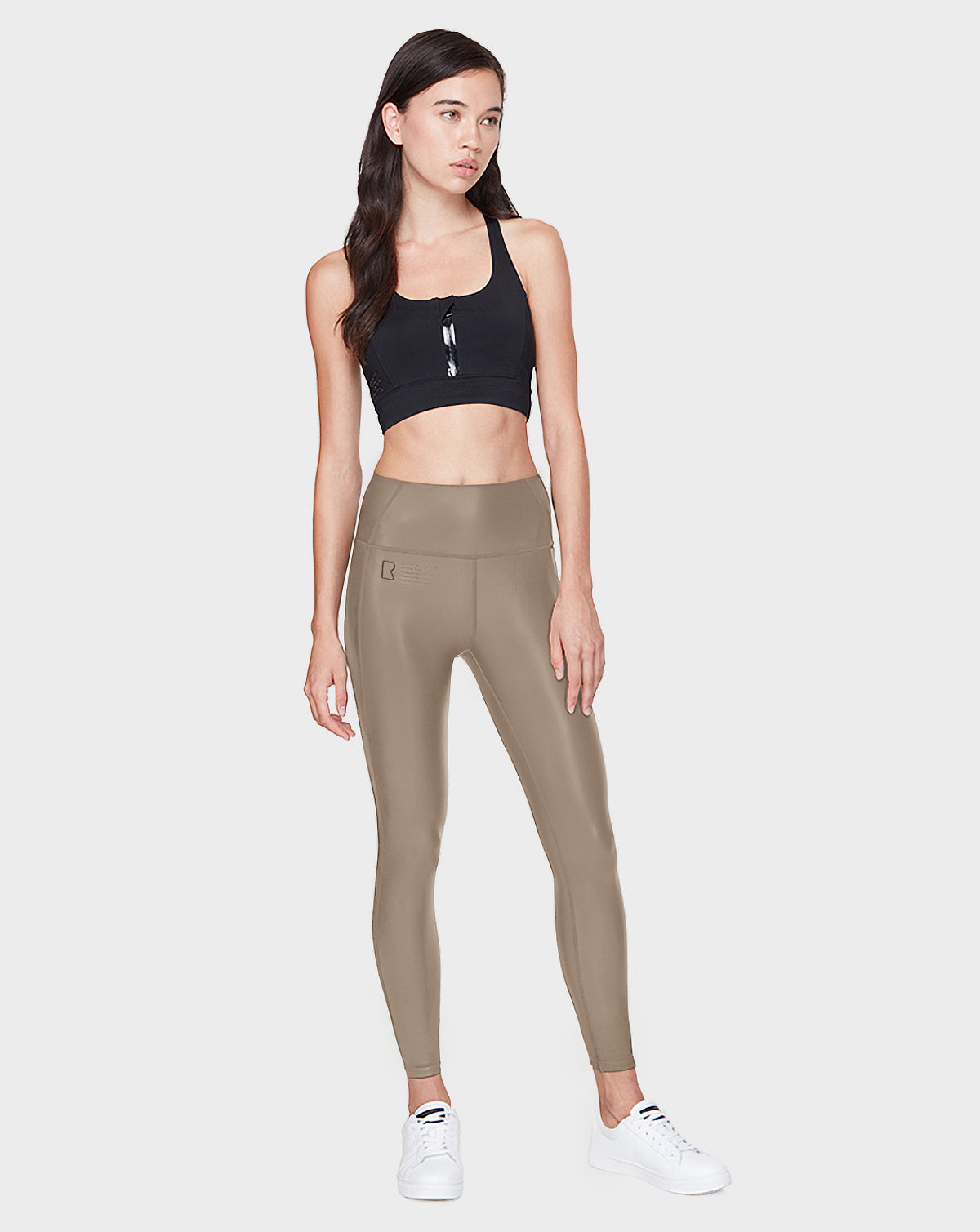 A woman with long, dark hair stands against a plain background wearing Rudsaks SHIRIN L - 8521034 Cement high-waisted leggings and a black sports bra, paired with white sneakers. She looks slightly to the side with a neutral expression.
