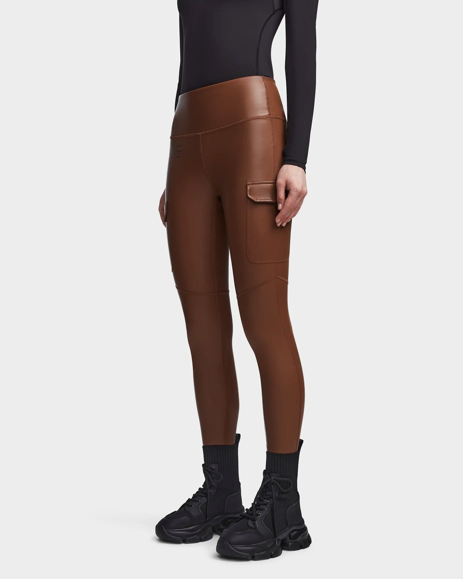A person wears Rudsak’s SIEN L - 8521042 COHIBA brown vegan leather leggings with a side pocket, styled with a black long-sleeve top, socks, and chunky sneakers against a white background—ideal for highlighting activewear essentials.