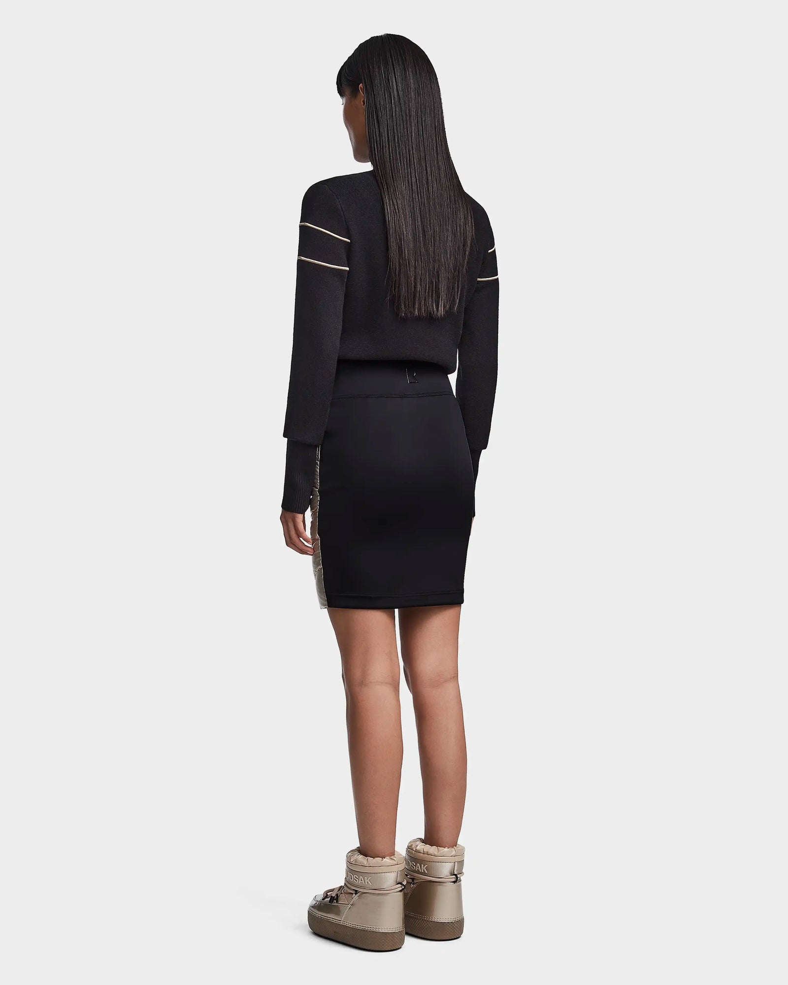 A woman with long straight hair stands facing away, wearing the Rudsak MARZI - 8523528 Metallic Ivory skirt, a black long-sleeve top with striped sleeves, and beige fur-lined ankle boots against a white background.