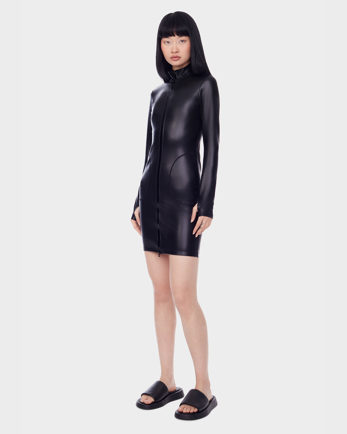 A woman with straight black hair wears the SHANA - 8522508 BLACK fitted zip-up stretch vegan leather dress by Rudsak and black slides, standing against a plain white background.