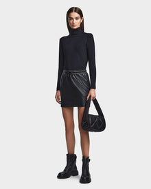 A woman with straight, dark hair and a neutral expression stands against a plain background wearing the RUDSAK PORTIA - 8523033 BLACK turtleneck, a vegan leather mini skirt, black ankle boots, and holding a black quilted handbag.