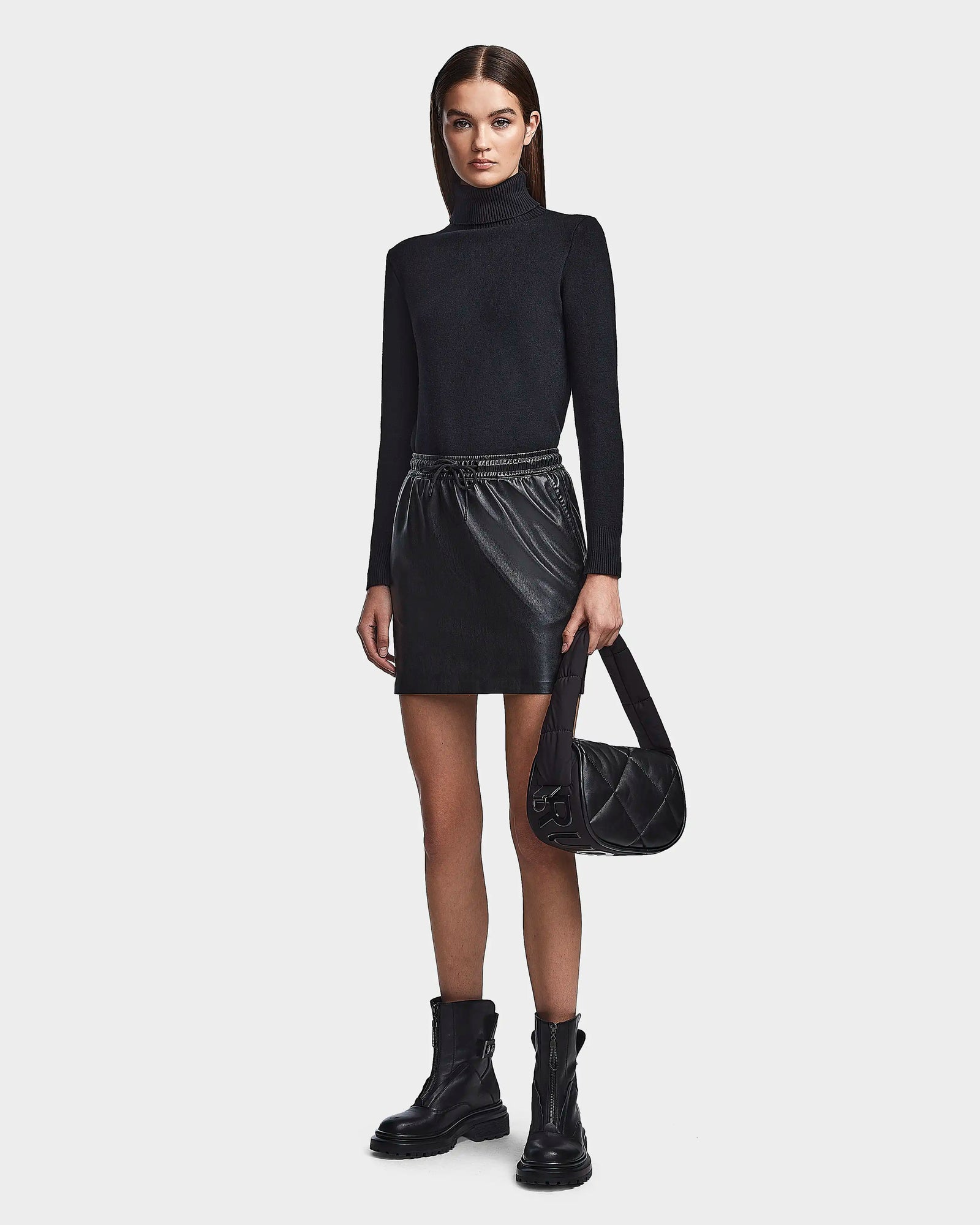 A woman with straight, dark hair and a neutral expression stands against a plain background wearing the RUDSAK PORTIA - 8523033 BLACK turtleneck, a vegan leather mini skirt, black ankle boots, and holding a black quilted handbag.