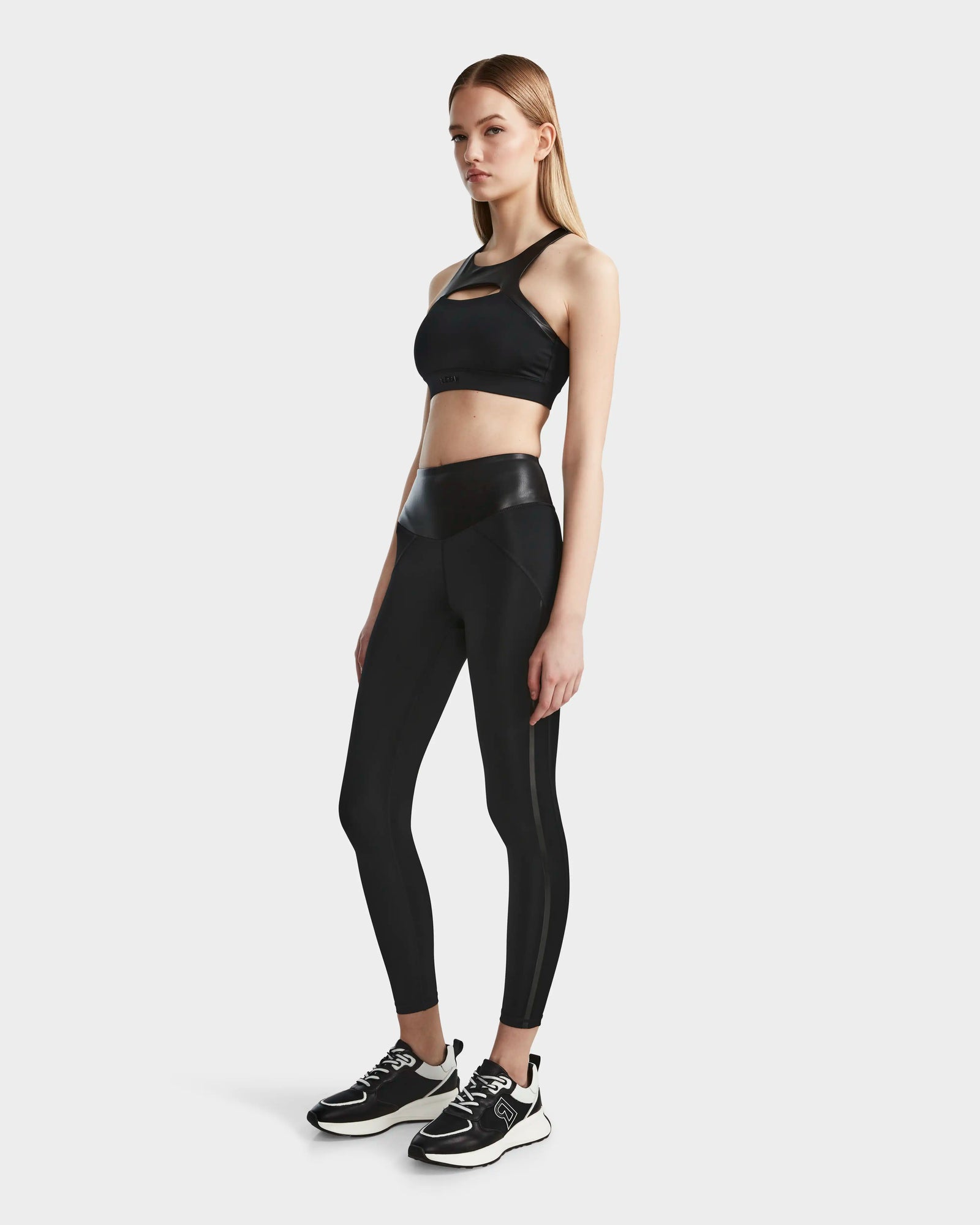 A woman poses in the ARYA - 8523002 BLACK sports bra and matching high-waisted leggings by RUDSAK, paired with black sneakers with white soles, set against a plain light background.