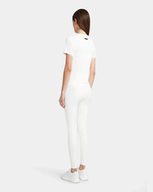 A woman with straight brown hair stands facing away on a light gray background, wearing the Rudsak SUMI - 8524030 Cotton White womens essential tee, paired with white leggings and white sneakers.