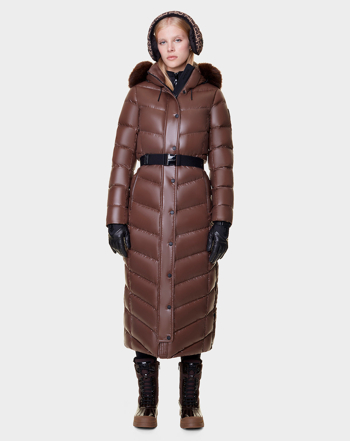 A woman faces forward in the Rudsak ELSIE - L121525 CAOCAO womens maxi down puffer with RDS-certified down, brown quilted design, fur-trimmed removable hood, black gloves and belt, lace-up boots, and earmuffs against a white background.