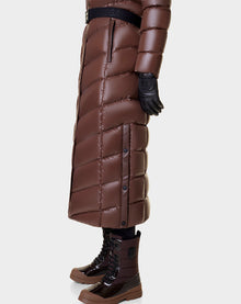 A person wears the Rudsak ELSIE - L121525 CAOCAO, a long brown womens maxi down puffer with black side buttons and a black waist belt, paired with black gloves and brown lace-up boots. The coat is filled with RDS-certified down; background is plain and light.