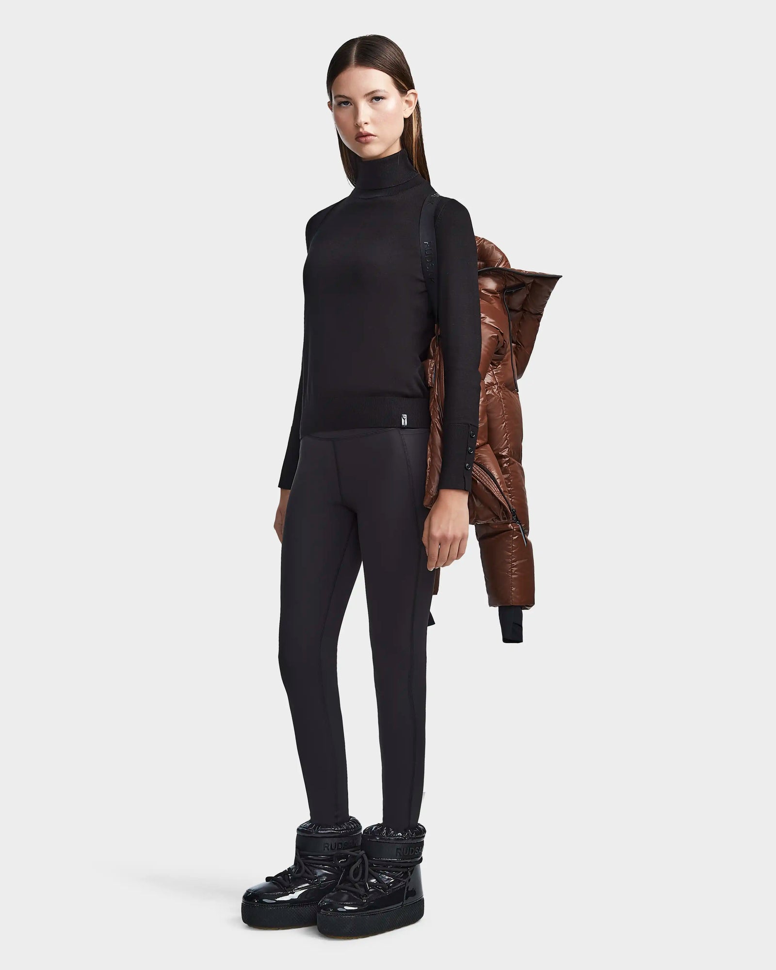 A woman with straight brown hair and a neutral expression stands facing forward in black leggings, boots, and a fitted turtleneck. The Rudsak RITA S - L123527 Cohiba weather-resistant puffer jacket is draped over her left shoulder.