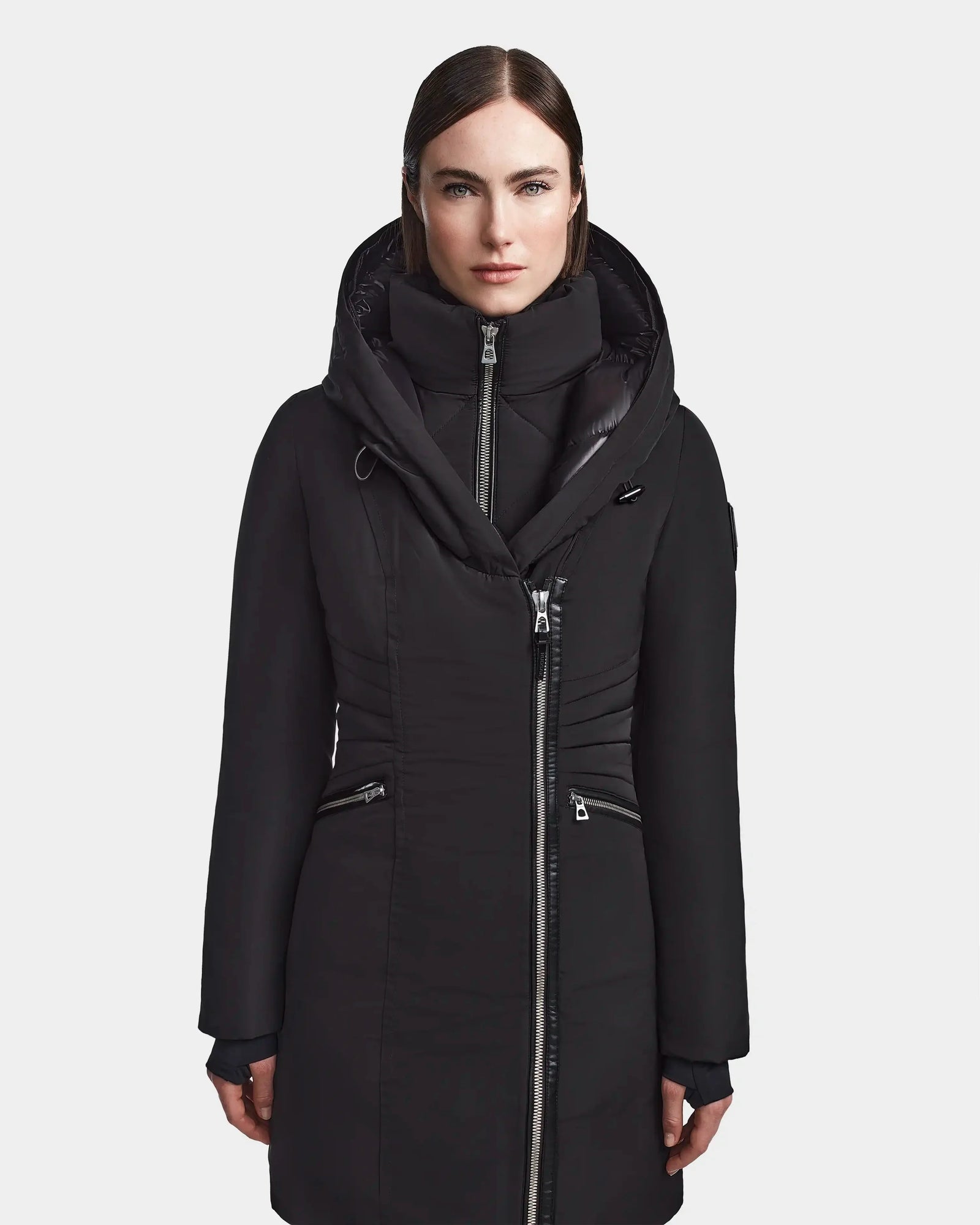 A woman with straight dark hair wears the Rudsak CORA - L123538 Black fitted parka, featuring multiple zippers, standing facing forward against a plain light background.