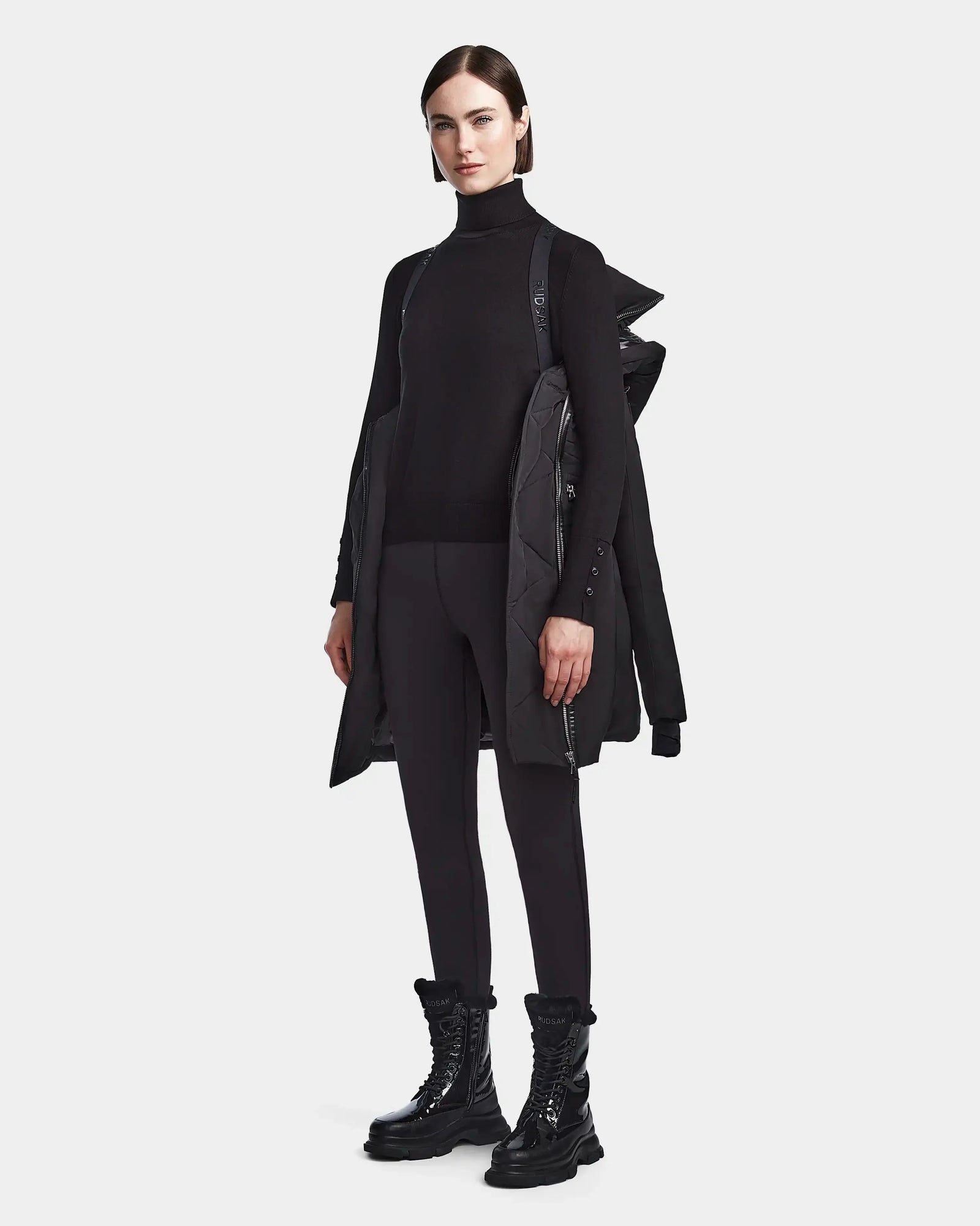 A woman poses against a white backdrop wearing the Rudsak CORA - L123538 Black sustainable winter coat draped over her shoulders, paired with a black turtleneck, leggings, and lace-up boots.