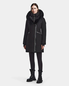A woman faces forward in the Rudsak CORA - L123538 Black parka, a long sustainable coat with a fur-trimmed collar, high neck, and silver zippers. She pairs it with black leggings and lace-up boots against a plain light gray background.