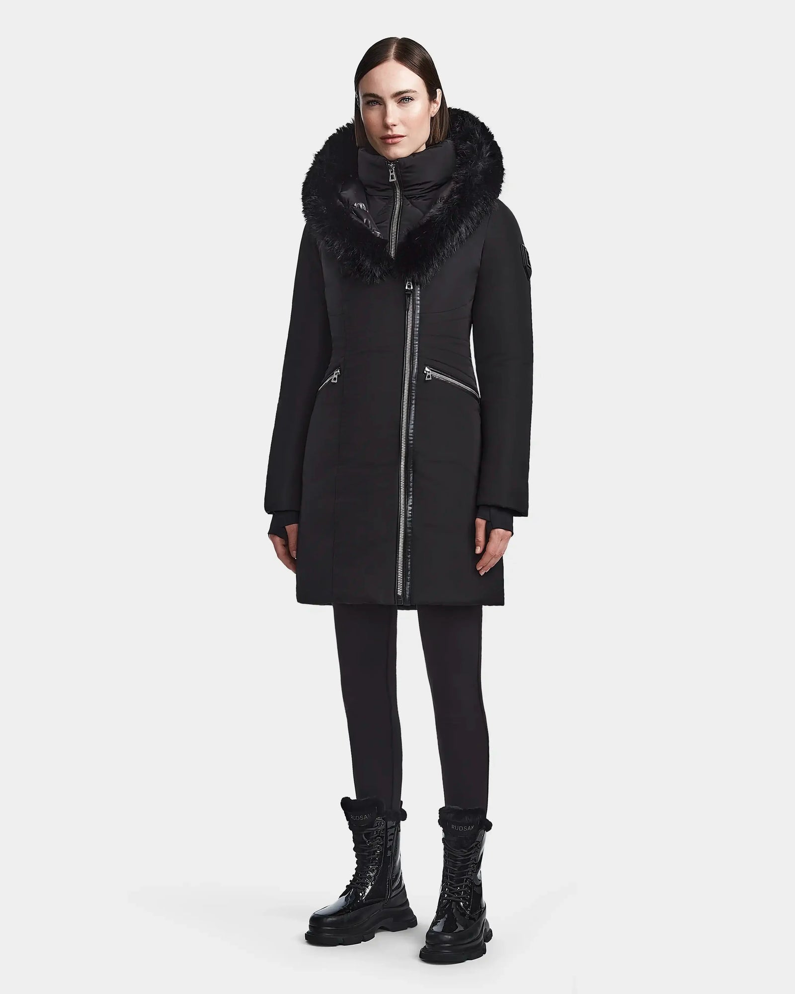 A woman faces forward in the Rudsak CORA - L123538 Black parka, a long sustainable coat with a fur-trimmed collar, high neck, and silver zippers. She pairs it with black leggings and lace-up boots against a plain light gray background.