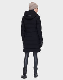 A person wearing the Rudsak ELISA - L121512 BLACK womens quilted down puffer, along with black leggings, boots, gloves, and a beige knit beanie stands facing away against a plain light background.
