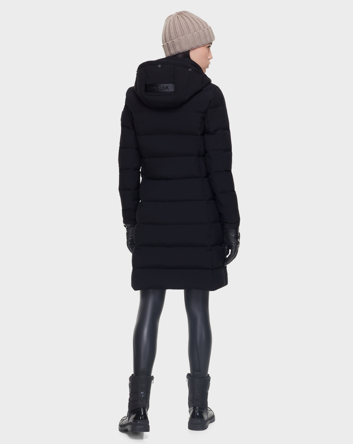 A person wearing the Rudsak ELISA - L121512 BLACK womens quilted down puffer, along with black leggings, boots, gloves, and a beige knit beanie stands facing away against a plain light background.